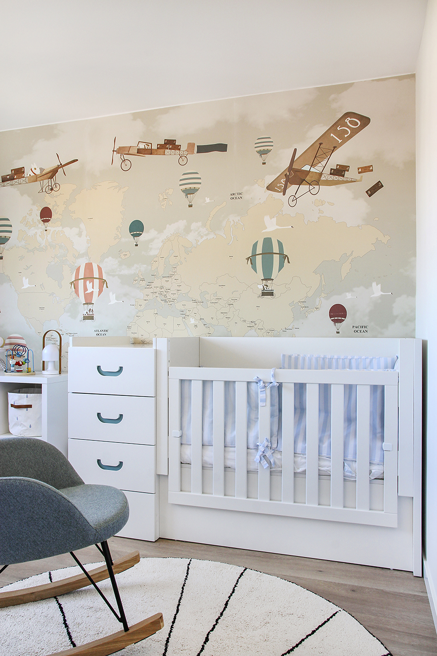 Reforma integral y decoración vivienda. Habitación de bebé con pared principal empapelada con mapamundi y avionetas. Mueble cuna cambiador y sillón mecedora sobre alfombra circular.